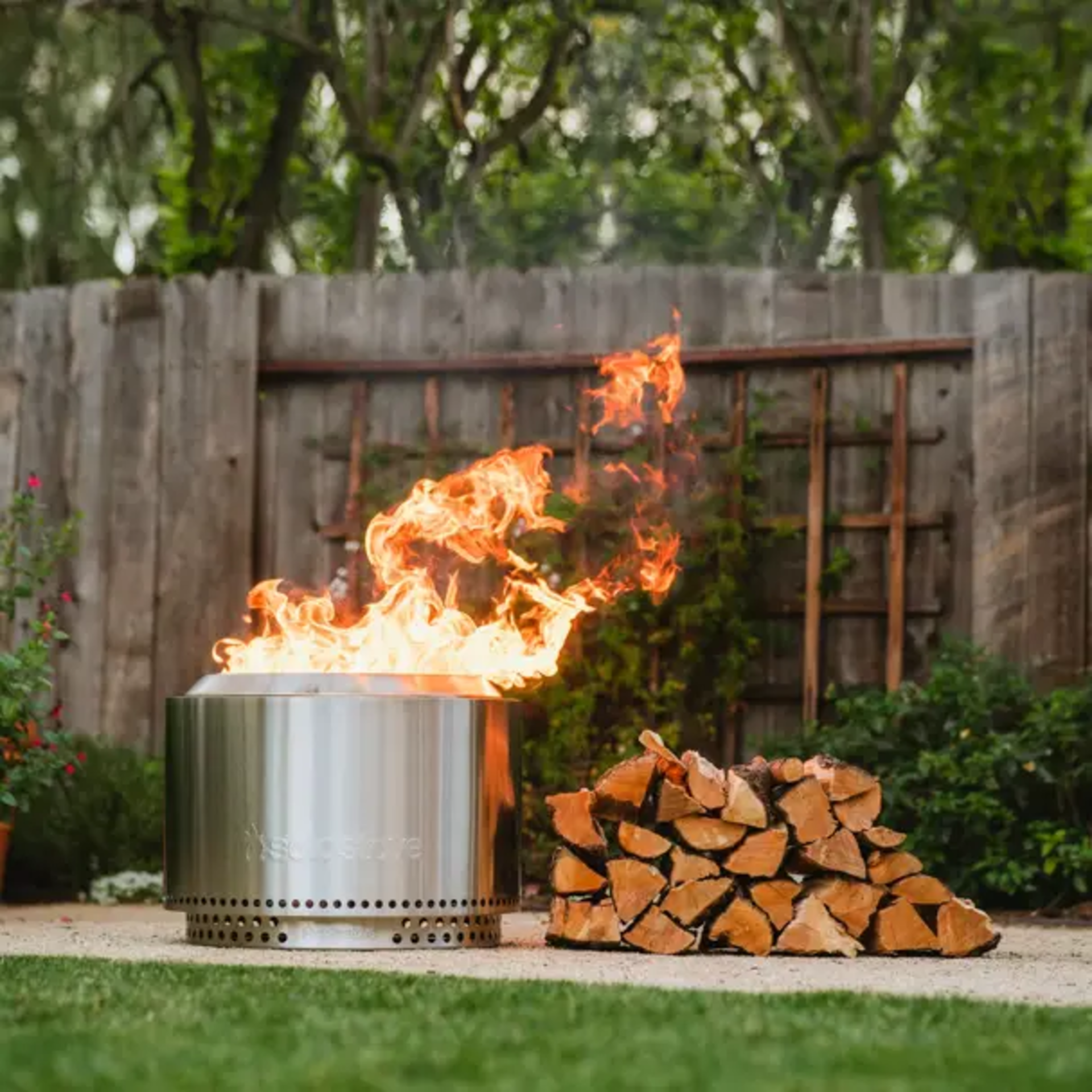 Rauchfreie Edelstahl-Feuerstelle Modell Solo Stove Yukon mit kräftigem Holzfeuer im Garten – modernes Design für gemütliche Abende im Freien, inklusive Brennholzstapel.