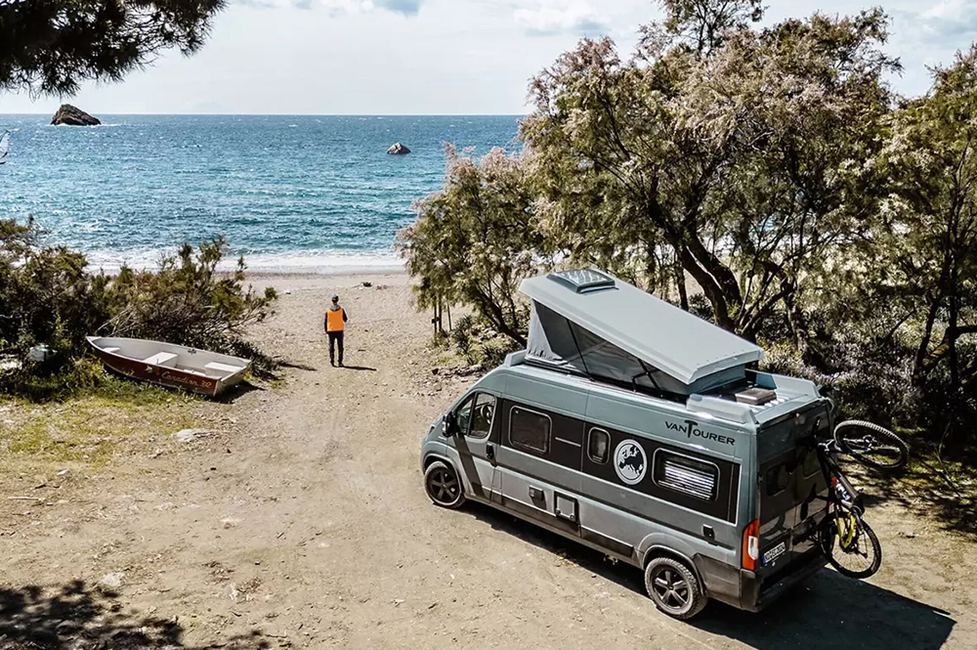 Ein blauer Camper Van von VANTourer steht an einem Meer in einer Dünenlandschaft mit Bäumen.
