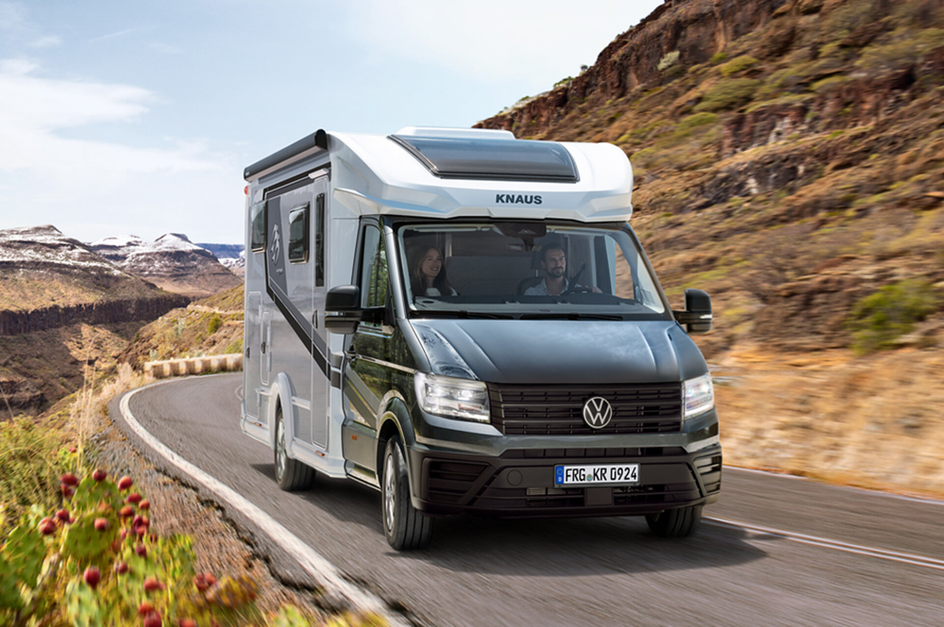 A KNAUS motorhome drives along a road through a mountain landscape.