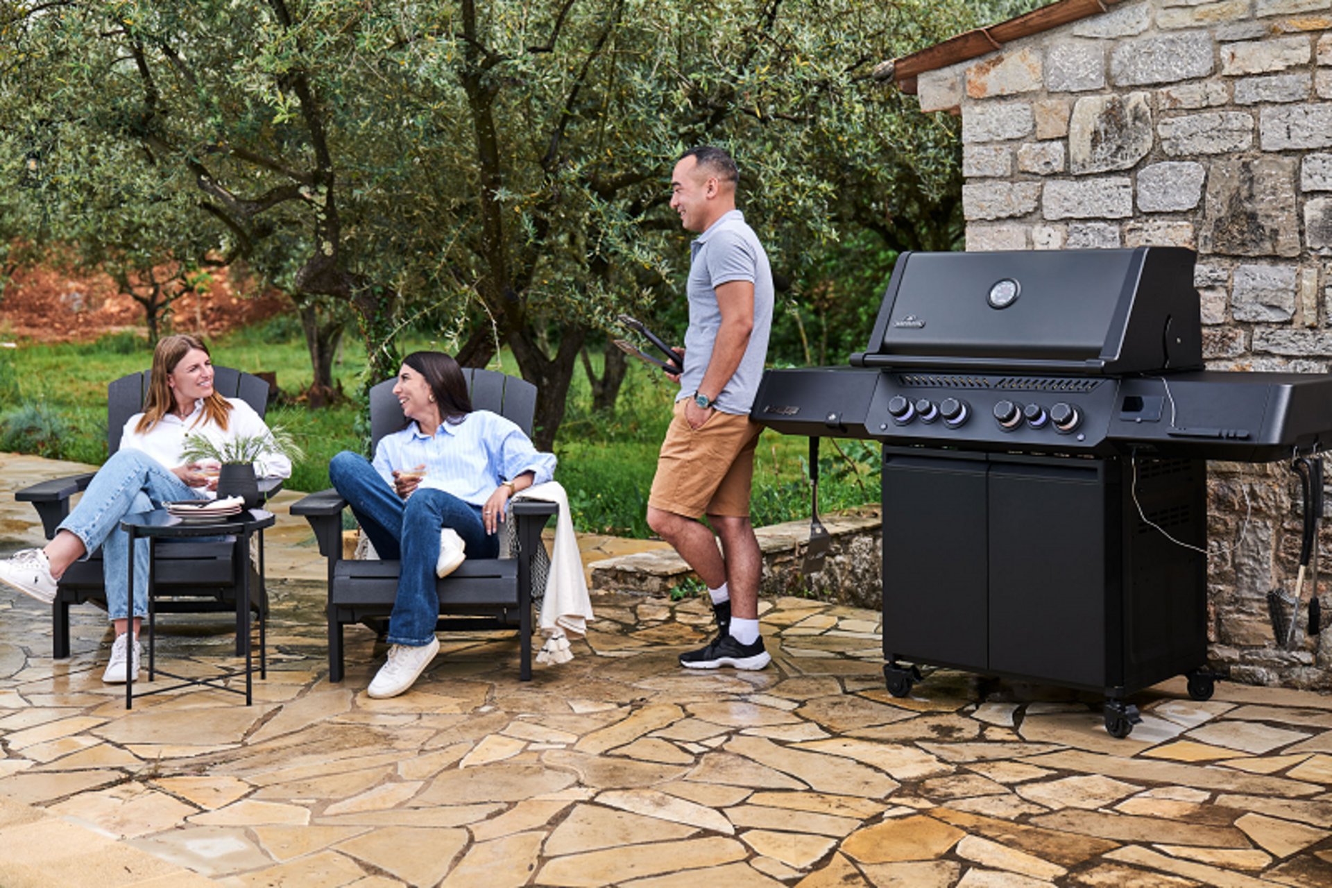 2 Frauen sitzen unter freiem Himmel und unterhalten sich mit einem Mann der an einem Napoleon Gasgrill Modell Phantom Prestige 500 Connected steht.