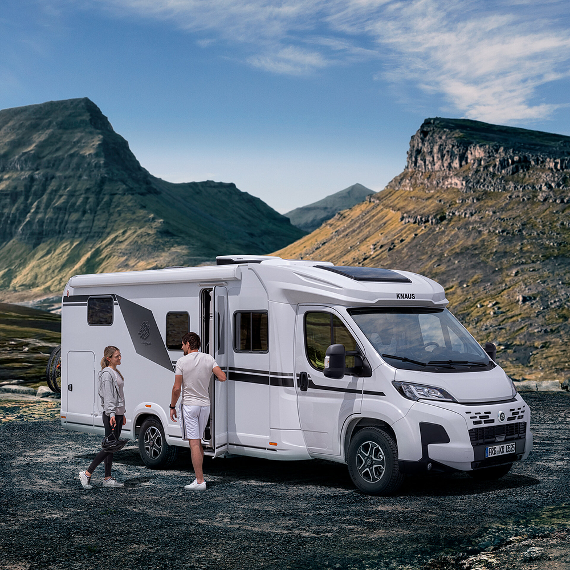 Ein weißes Wohnmobil steht in einer Berglandschaft. Zwei Personen stehen davor.