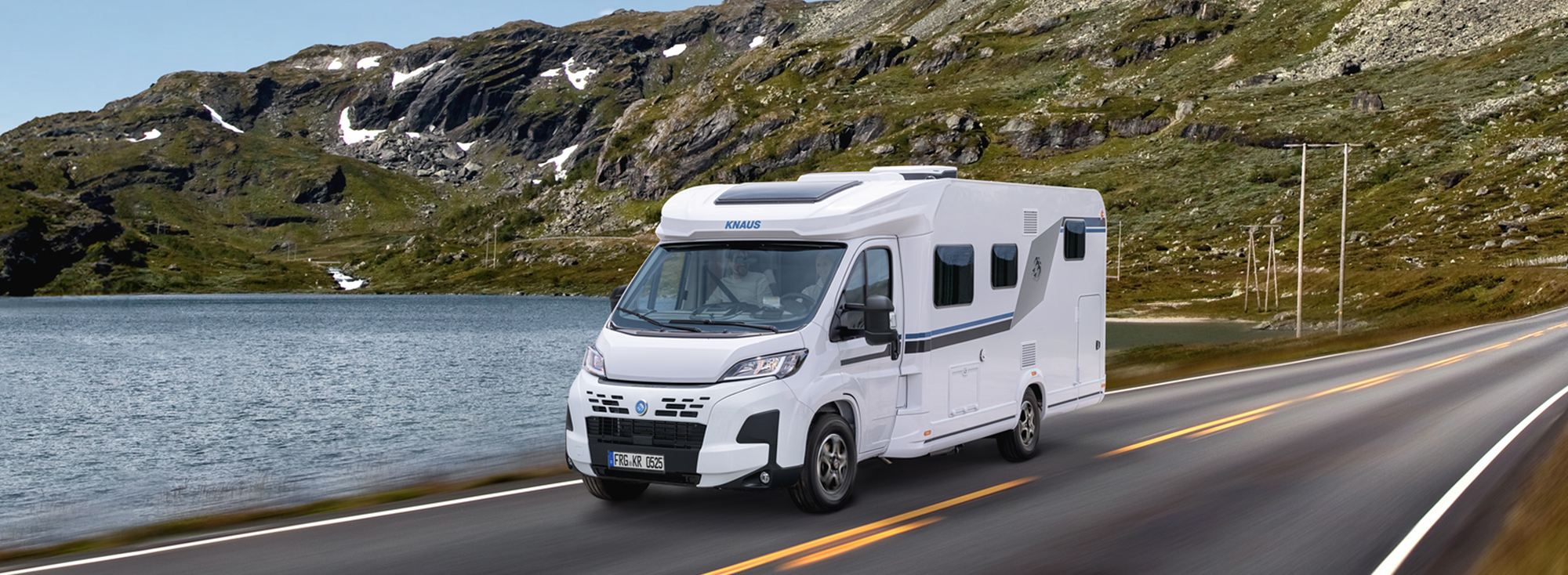 A KNAUS L!VE TI drives along a road next to a lake.