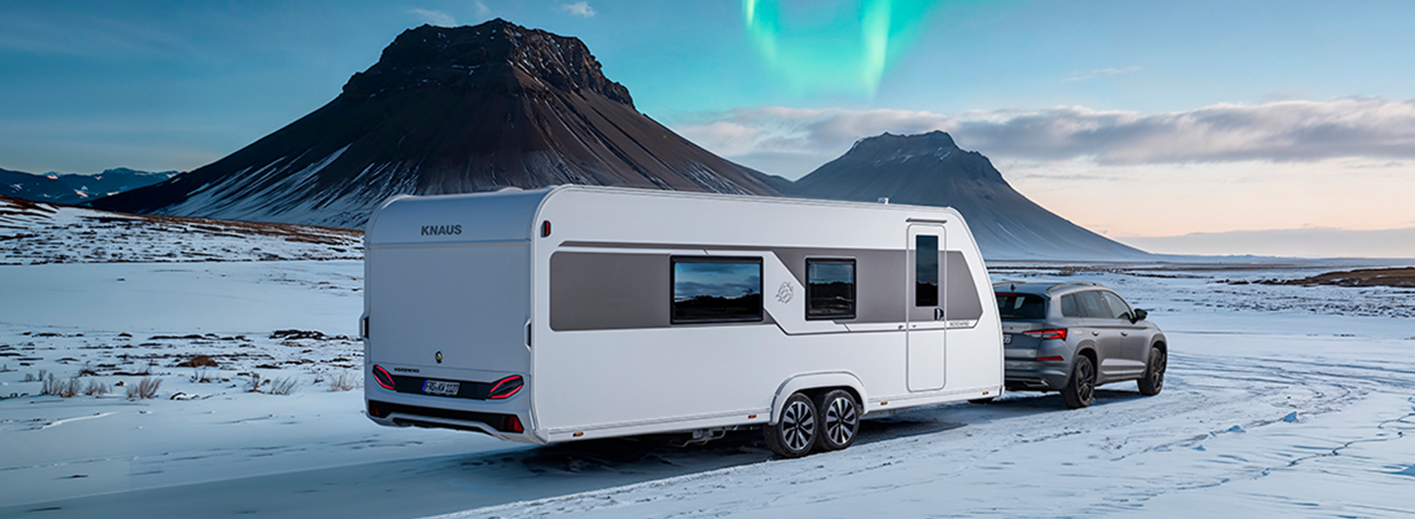 A white caravan on snow with the northern lights in the sky.