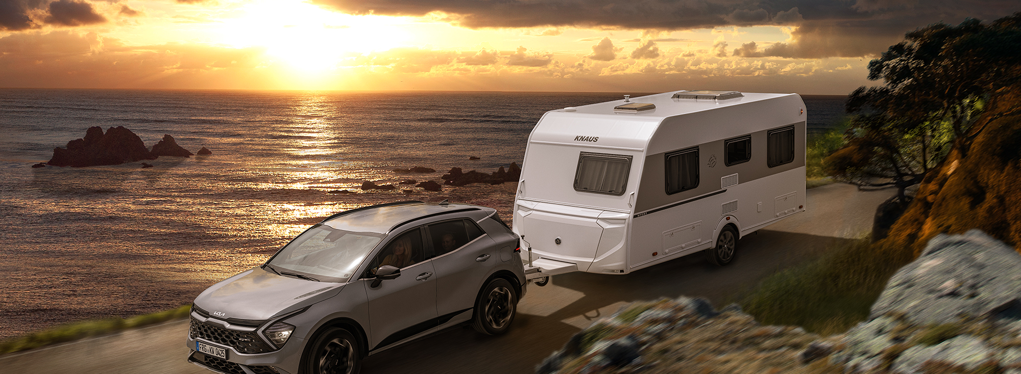 Een witte caravan wordt door een grijze SUV langs de zee getrokken.